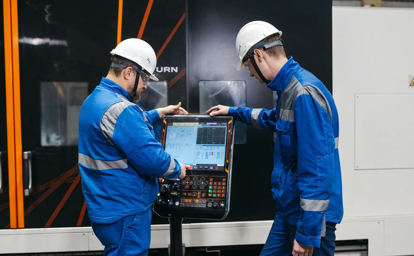 Zwei Techniker mit Schutzhelm prüfen ein Programm am Bedienpult einer CNC-Maschine