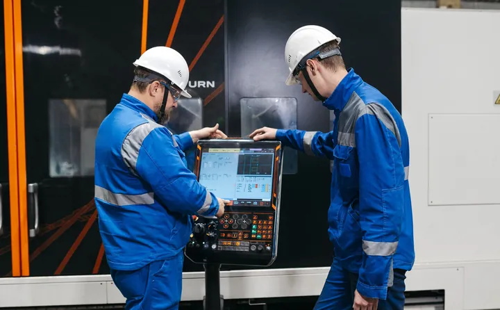 Zwei Techniker mit Schutzhelm prüfen ein Programm am Bedienpult einer CNC-Maschine